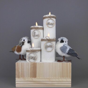 Four flameless pillar candles wrapped in white crochet sleeves with jute twine and round wooden tags, displayed on a wooden block with two decorative plush birds.