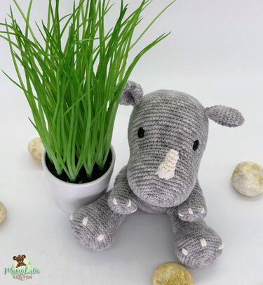 Graues, sitzendes Amigurumi‑Nashorn mit kleinem weißem Horn und schwarzen Augen, handgehäkelt, neben einer Topfpflanze auf weißem Untergrund.