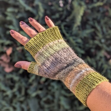 Hand mit gestrickter, fingerloser Armstulpe in Grün-, Grau- und Beigetönen vor unscharfem Hintergrund.