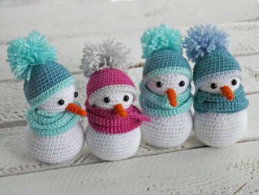 Reihe von vier gehäkelten Schneemännern mit bunten Mützen, Pompons und Schals auf hellem Holzuntergrund.