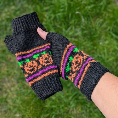 Schwarze, gestrickte fingerlose Handschuhe mit orangefarbenen Kürbissen und bunten Streifen, an einer Hand getragen