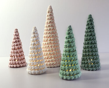 Five crocheted cone trees in pink, cream, light green, and dark green, softly lit from the inside on a white surface.