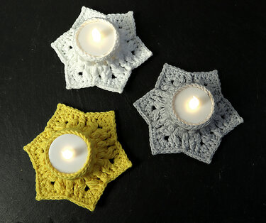 Three crocheted six-point star tea light coasters in white, gray and yellow holding lit tea lights on a dark surface