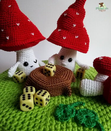 Drei gehäkelte Pilze mit roten Häubchen, braunem gehäkeltem Baumstumpf, gelben Würfeln und gehäkeltem Kleeblatt auf grüner Unterlage
