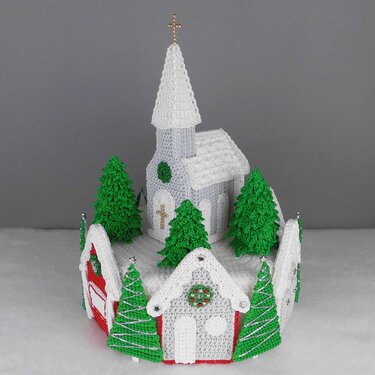 Crocheted miniature church with steepled roof and pine trees on a snowy round base