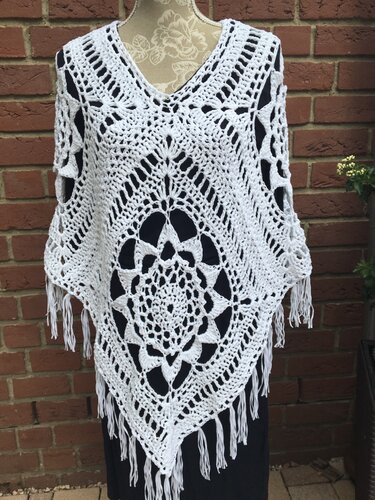 Weißer gehäkelter Damenponcho mit zentralem Mandala, V-Ausschnitt und Fransen auf Schneiderpuppe vor Backsteinwand