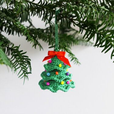 Gehäkelter grüner Weihnachtsbaum-Anhänger mit roter Schleife und bunten Perlen an einem Tannenzweig