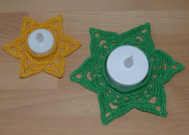 Zwei sternförmige gehäkelte Untersetzer in Grün und Gelb mit weißen Teelichtern auf Holztisch