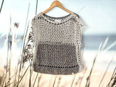 Gehäkeltes Damen‑Shirt in Creme und Braun mit Netzmuster an einem Holzbügel vor Strandkulisse.