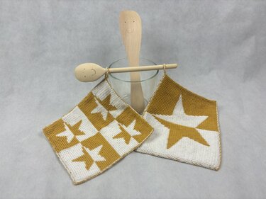 Two knitted square potholders with mustard yellow and off-white star motifs, displayed with a glass holding two wooden spoons.