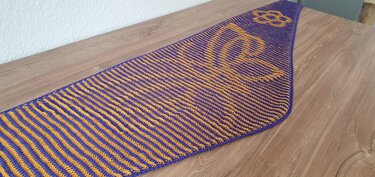 Triangular knitted shawl laid flat with narrow purple and mustard-yellow stripes and a yellow motif at the narrow end