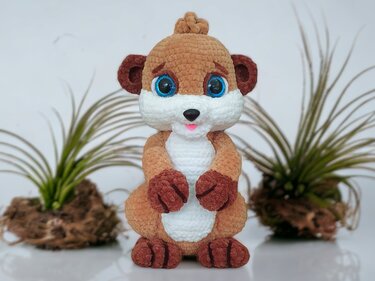 Hand-crocheted brown and white meerkat amigurumi with blue safety eyes and a small pink mouth, standing upright in front of blurred plants.