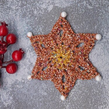 Gehäkelter sechszackiger Stern aus rotbraunem Garn mit weißen Perlen an den Spitzen, daneben rote Dekobeeren auf grauem Untergrund.