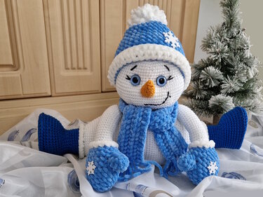 Gehäkelter Schneemann in Blau und Weiß mit Mütze, Schal und Fäustlingen neben einem kleinen Tannenbaum.