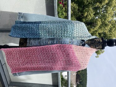Zwei gehäkelte Loop-Schals mit Farbverlauf (Blau-Grün und Rosa) auf einer Puppe vor einem Geländer und Bäumen