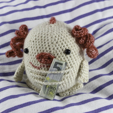 Rundes, weiß-beiges gehäkeltes Amigurumi mit rotbraunen Locken und einem gefalteten 5-Euro-Schein.