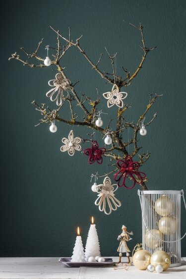 Natürlicher Zweig mit creme- und bordeauxfarbenen gehäkelten Blüten-, Stern- und Engelsanhängern, kleinen Perlenkugeln, goldenen Christbaumkugeln und zwei weißen Tannenbaumkerzen auf einem Tisch.