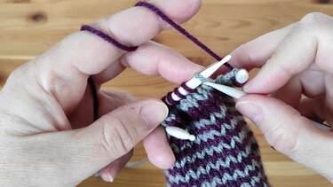 Nahaufnahme von Händen beim Stricken mit Doppelspitzen, mit violetter und grauer Wolle