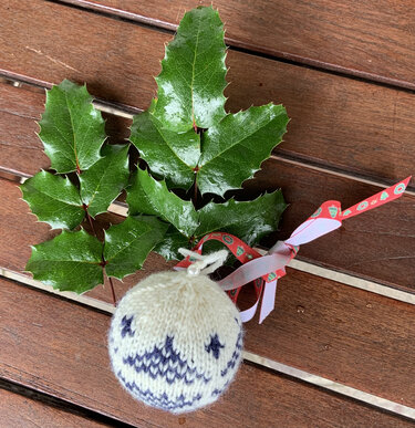 Weiße gestrickte Kugel mit blauem Muster und Schleife auf Holz neben grünen Stechpalmenblättern