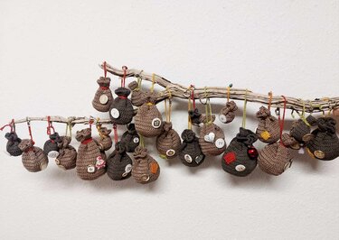 Gehäkelter Adventskalender mit 24 nummerierten Säckchen an Ästen vor weißer Wand