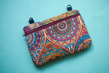 Rechteckige Stofftasche mit buntem Mandala-Muster, Reißverschluss und zwei D-Ringen auf hellblauem Hintergrund