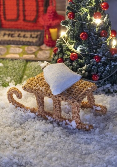 Gehäkelter Mini-Schlitten mit weißem Kissen vor beleuchtetem Weihnachtsbaum auf Kunstschnee