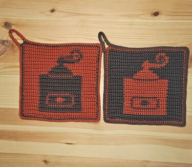 Zwei gehäkelte quadratische Topflappen mit Kaffeemühlen-Motiv in invertierten Farben auf Holzuntergrund