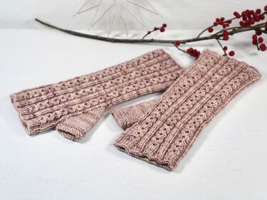 Zwei gestrickte Armstulpen in Rosé mit Lochmuster auf weißem Hintergrund, dekoriert mit einem Beerenzweig.