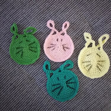 Vier gehäkelte Untersetzer in Pastellrosa, Gelb und zwei Grüntönen mit Öhrchen, arrangiert auf dunklem Stoff