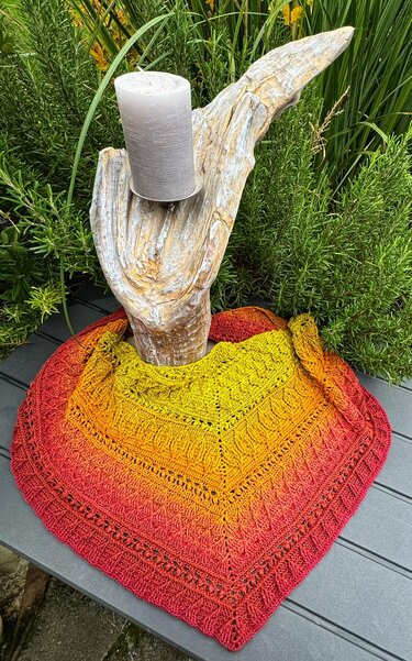 Handmade crochet triangle shawl in yellow-to-red gradient draped around a driftwood candle holder on a gray patio table