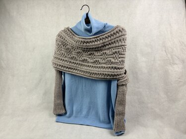 Grau gestrickter Schulterschal mit Relief- und Zopfmuster über hellblauem Pullover an Bügel