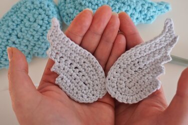 Pair of small light-gray crochet wings held in hands with blue crochet in the background.