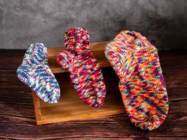 Drei handgestrickte, bunte Socken in aufsteigender Größe, drapiert auf einer Holzkiste vor neutralem Hintergrund.