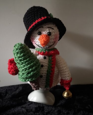 Weißer gehäkelter Schneemann-Eierwärmer mit schwarzem Zylinder, rotem Schal, grüner Tanne und Glocke auf einem Eierbecher