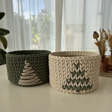 Zwei runde gehäkelte Körbe in Dunkelgrün und Natur mit Tannenbaum‑Motiven auf einem Tisch vor hellen Gardinen