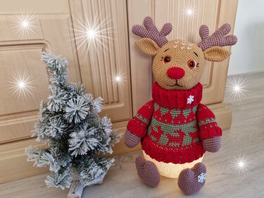 Gehäkeltes Rentier-Amigurumi mit rotem Pullover, Geweih und leuchtendem Bauch neben einem kleinen beschneiten Weihnachtsbaum