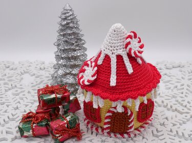 Gehäkeltes Lebkuchenhaus in Rot und Weiß mit Zuckerstangen-Akzenten, kleinen Geschenkpaketen und silbernem Baum auf weißem Schneeflocken-Untergrund