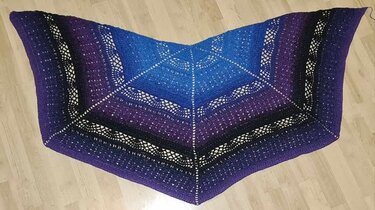 Triangular crochet shawl with blue-to-purple ombre and lace bands, laid flat on a light wooden floor.