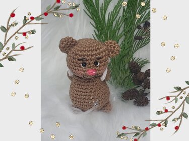 Kleiner brauner Amigurumi-Teddybär mit Schneeflocke und rosa Schnauze, umgeben von Tannenzweigen und Zapfen.