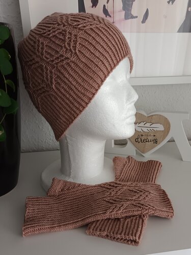 Roséfarbenes gestricktes Mützen- und Stulpen-Set mit Zopfmuster; Mütze auf Schaufensterkopf, Stulpen liegend.