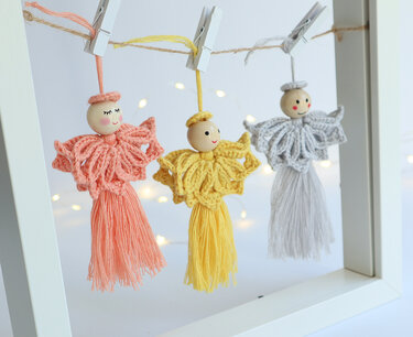 Three crochet angel ornaments with wooden bead heads and tassel skirts in coral, yellow and gray hanging on twine