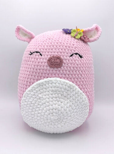 Rosa Amigurumi-Kuscheltier mit weißem Bauch, geschlossenen Augen, kleinen Ohren und bunten gehäkelten Blumen am Kopf.