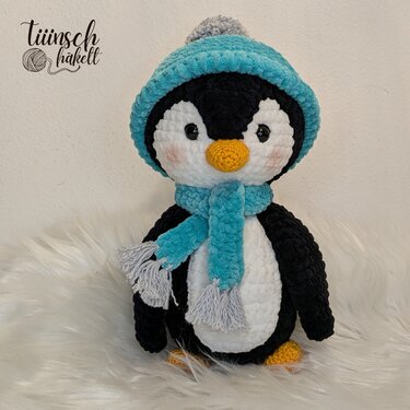 Gehäkelter Amigurumi-Pinguin mit türkiser Mütze und Schal auf hellem flauschigem Untergrund