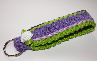 Gehäkelter Schlüsselanhänger in Lila und Limettengrün mit weißer Stoffrose und Metallring auf weißem Hintergrund