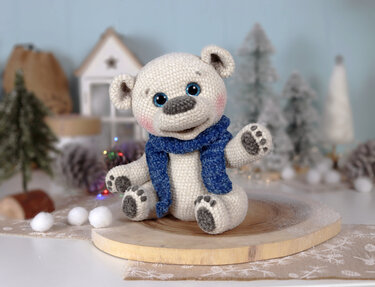 Gehäkelter weißer Amigurumi-Eisbär mit blauem Schal auf Holzscheibe vor winterlicher Dekoration