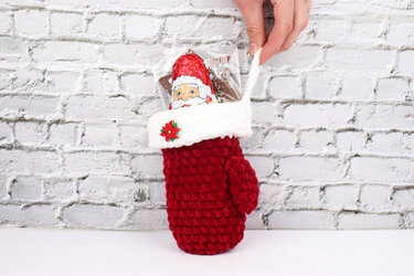 Red crocheted mitten treat holder with white cuff and poinsettia holding a foil-wrapped Santa chocolate and a star candy.
