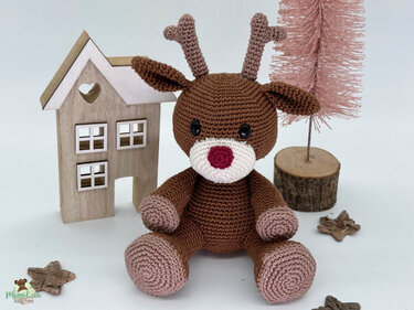 Braunes gehäkeltes Amigurumi-Rentier mit roter Nase vor Holzhaus und rosafarbenem Deko-Baum.