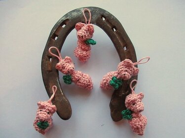 Fünf rosa gehäkelte Glücksschweinchen mit grünen Kleeblättern an einem rustikalen Hufeisen.