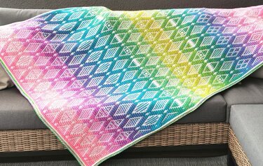 Gehäkelte Decke mit Regenbogen-Farbverlauf und weißem Rautenmuster auf grauem Sofa
