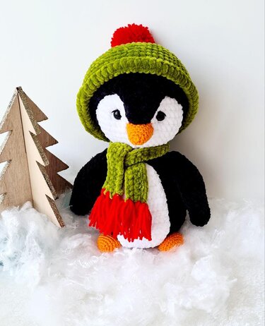 Gehäkelter Amigurumi-Pinguin mit grüner Mütze, rotem Bommel und grünem Schal, orangefarbener Schnabel und Füße